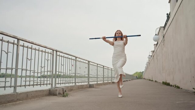 Woman in white dress walks along outdoor pathway holding blue pole with water and metal railings visible beside her. Woman maintains steady movement through scene under soft daylight