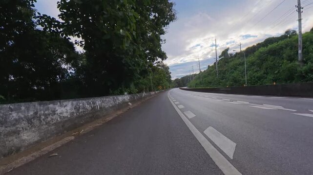 Scenic winding road through lush green forest with concrete barriers, chevron signs, utility poles and power lines under a partly cloudy sky.
