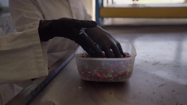 A close-up shows Latinx scientists, a man and a woman, inspecting colorful shredded HDPE plastic particles in a container on a steel table, preparing them for recycling into sustainable construction m