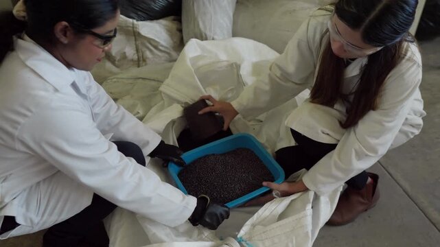 Two female scientists in lab coats, safety glasses, and gloves, examining and transferring shredded HDPE plastic from a large bag into a smaller container for ecological construction material recyclin