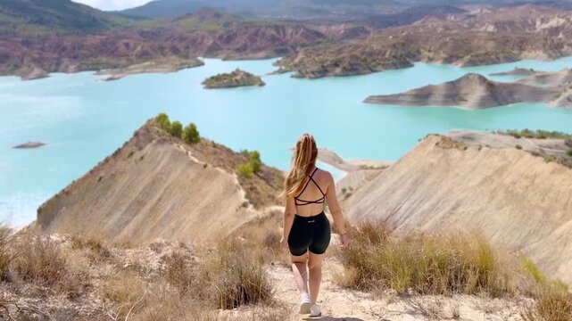 Cinematic Rear View of Woman in Blue Dress Walking Across Desert Hills Toward Turquoise Lake at Barranco de Gebas Murcia Spain