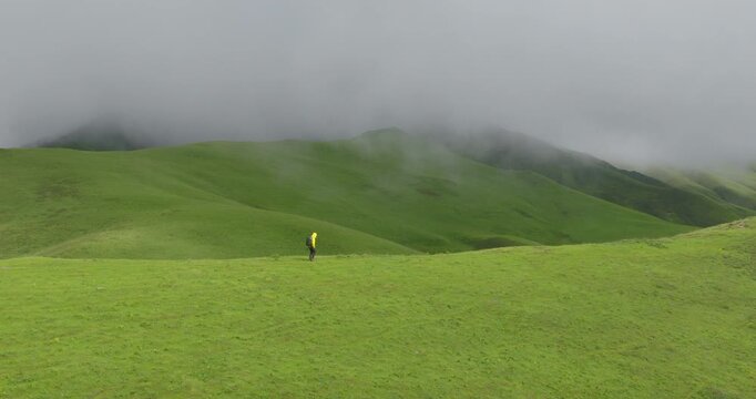 Drone pushes in towards a lone hiker in a yellow jacket in Badimalika, Bajura, Nepal, trekking across lush green grassland valley under heavy monsoon clouds fogging up the scenery