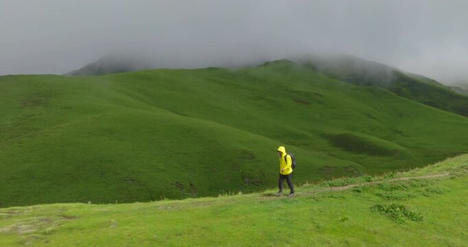 Badimalika, Bajura, Nepal, aerial pull out shot from a lone hiker in a yellow jacket trekking along a ridge to reveal vast, fog-shrouded green pastures and mountain slopes.