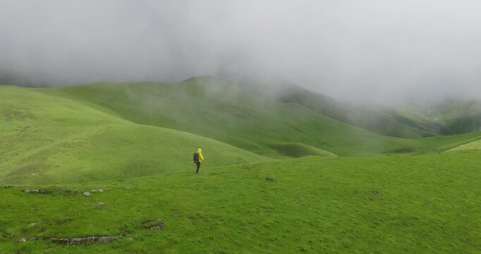 Badimalika, Bajura Nepal, drone shot following a lone hiker in a yellow jacket trekking along sprawling green alpine pastures enveloped by low monsoon fog under cloudy skies