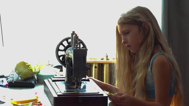 A medium static profile camera shot captures a young blonde girl sitting at a table removing fabric while taking apart a vintage mechanical sewing machine setup during a lesson in Budapest Hungary.