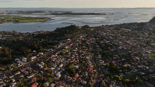 Panaromic view over Mount Roskill, Auckland, revealing suburban homes, greenery, streets, traffic, an ocean and warm evening sunlight across the neighborhood.