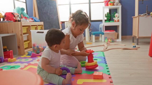 Toddler boy and toddler girl stacking colorful plastic rings on foam puzzle mat in a daycare building; playful joy.