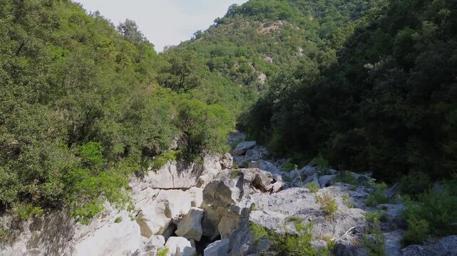 Drone travels along the rocky bed of Acheron River in Greece. Large pale boulders and narrow channels lie between densely wooded slopes, with patches of sunlight and shade.
