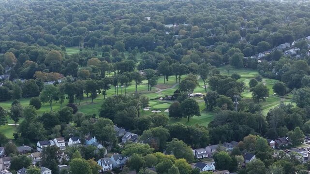 A drone glides across a green golf course bordered by mature trees and nearby homes, revealing curved fairways, bright sand bunkers and patches of morning light across a wooded suburban landscape.