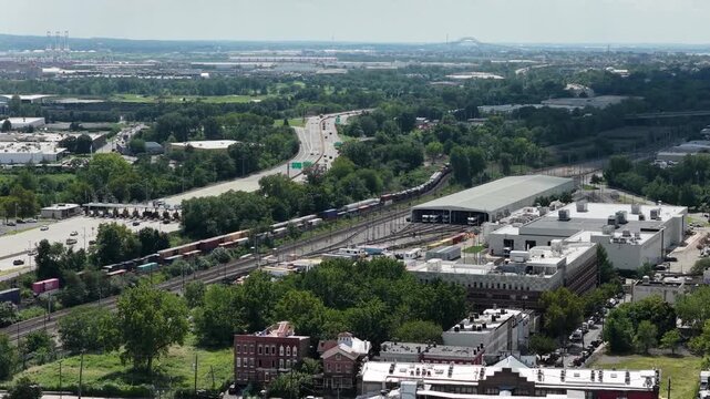 A drone moves toward a rail yard in Jersey City, NJ, revealing parallel tracks, colorful freight cars and long service buildings, with surrounding roads and dense green trees in summer daylight.