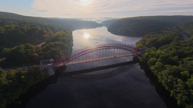 Aerial view facing the sun over New Croton Reservoir, surrounded by green trees. The camera dolly in and tilt down over the AMVETS bridge, ideal fro travel, environmental and cinematic B-roll content.