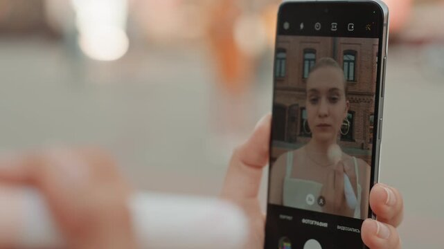 Woman applies makeup using brush while being photographed on smartphone. subject wears white top and necklace with background showing urban building exterior. Soft lighting creates natural