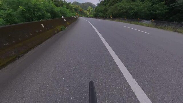 Cyclist POV riding on a winding mountain road through lush green forest, with guardrails, road signs, concrete barriers and distant mountains.