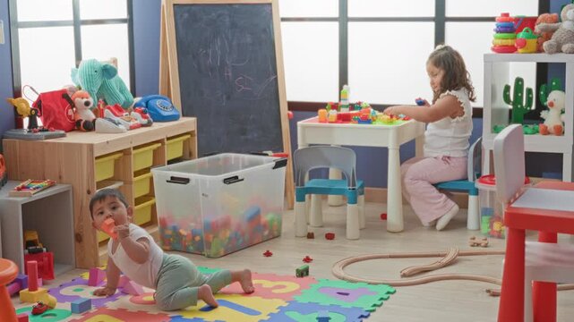 Toddler crawling on colorful foam mat chewing a block while girl at small table stacks plastic bricks beside storage bin and chalkboard in building; playful bonding.