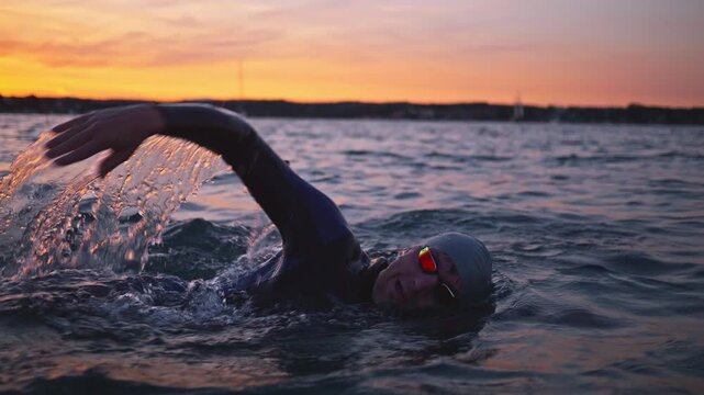 A young asian woman swims vigorously through ocean waves at sunset, her determination evident in every stroke. The scene represents sport training and the challenge of a triathlon.