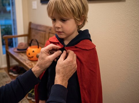 Parent helping young boy put on a red cape costume for Halloween at home.