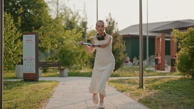 Woman in white dress practices tennis swings with imaginary ball on paved path through green park surroundings. Woman holds and moves blue racket dynamically demonstrating various strokes