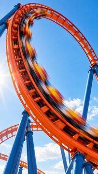 Orange roller coaster train speeding along elevated steel tracks against blue sky. Amusement park thrill ride and entertainment attraction. Theme park recreation.