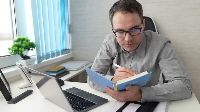 Focused professional taking notes with a pen in a notebook while looking at a computer screen. Business analysis, writing, and active work in the office. the teacher writes the curriculum.