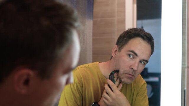 Young man shaving with an electric trimmer in front of bathroom mirror in the morning, following his daily grooming routine.