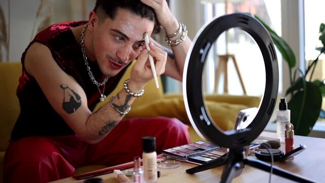 Young gender fluid person with tattoos and jewelry applying foundation with a makeup brush, sitting in front of a ring light and various cosmetic products for a beauty tutorial