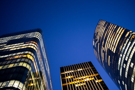 Illuminated modern high rise buildings with curved glass facades rise into a deep blue evening sky in a low angle view of contemporary urban architecture