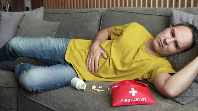 Sick man lying on sofa holding stomach, scattered pills and first aid kit nearby, health emergency.