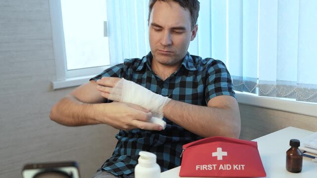 Man bandaging an injured hand at home while sitting at a table with a red first aid kit, providing himself with first aid after an injury.