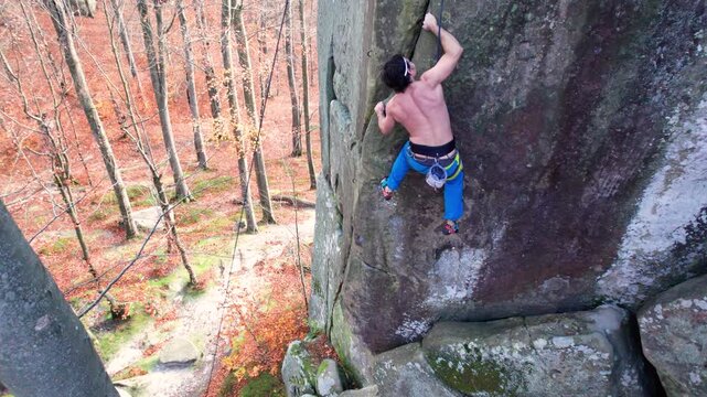 Shirtless athlete scales vertical rock formation in woodland, secured on climbing route. Drone footage for outdoor training, resilience, courage, and extreme sport storytelling