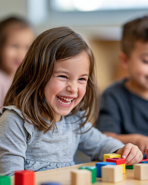 Happy young girl laughing while playing with colorful wooden building blocks at a bright preschool classroom table, experiencing joy and childhood development