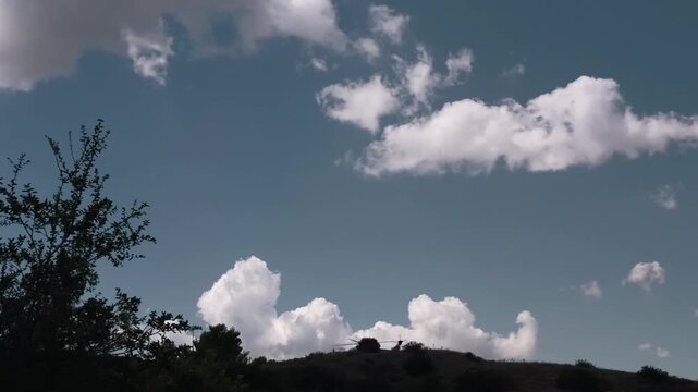 A firefighting helicopter waits on a ridge crest above Los Angeles with its rotors turning, silhouetted against towering cumulus clouds and dry chaparral brush along the hilltop.