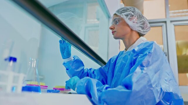 Young female laboratory technician in protective suit, glasses, and gloves, working with chemical reagents in a modern laboratory, researching vaccines and creating drugs for diseases