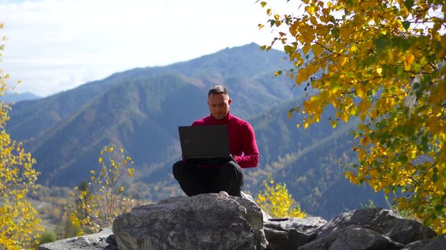 Focused man in a red sweater typing on a laptop during remote work on a beautiful mountain pass in autumn.