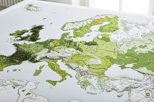 Three dimensional physical map of Europe with raised green terrain features representing forests wetlands and protected corridors on pale background under soft daylight