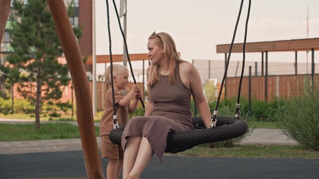 Woman and young boy interact at playground with child standing near her as woman sits on tire swing. scene takes place outdoors in urban park setting during daylight hours featuring greenery