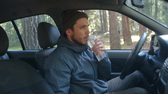 Young man wearing a beanie and jacket sitting in his car, drinking water from a reusable bottle to quench his thirst before starting the engine and continuing his road trip through the forest
