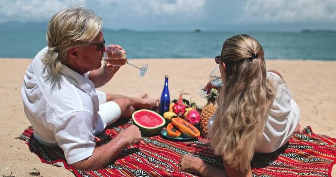 Senior couple relaxing on colorful blanket, enjoying beach picnic, drinking white wine, eating variety of tropical fruits on Samui Island, Thailand. Romantic scene, travel, holiday, vacation