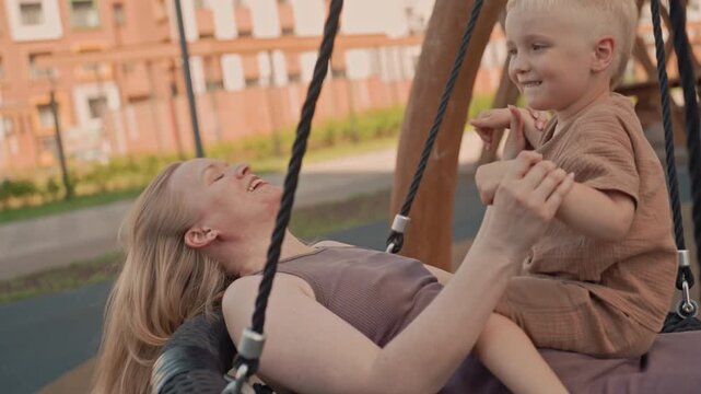 Woman and young boy share joyful moment on swing in outdoor playground. child interacts with adult creating intimate connection amidst soft natural light. Background reveals urban buildings