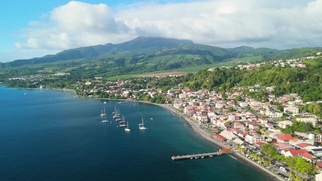 Aerial parallax shot of historic Saint Pierre town with red roof houses pier and anchored sailboats in Caribbean bay below cloud capped Mount Pelee volcano in Martinique