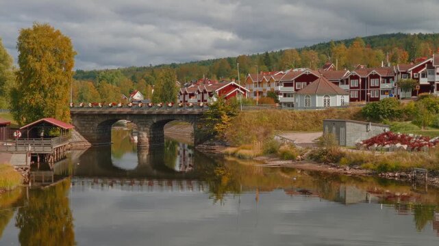 Static view of a charming Swedish town with traditional red houses and an old arched bridge reflected in calm water, surrounded by golden autumn trees and a pine forest hill. Mora, R&auml;ttvik, Sweden