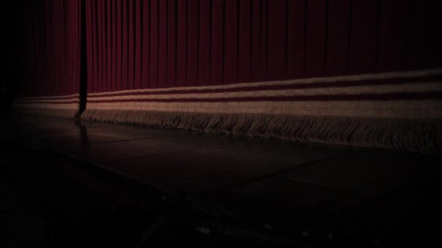 A dimly lit theater stage featuring a closed red velvet curtain with a fringed hem resting on a dark wooden floor, creating a dramatic and anticipatory mood.
