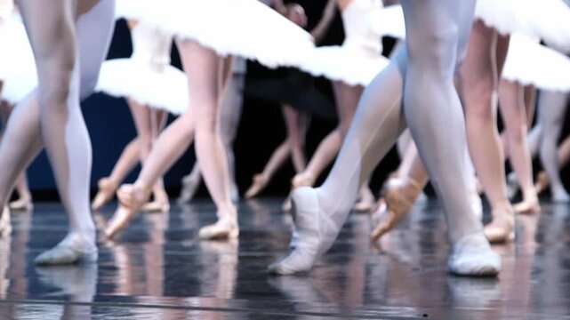 Low-angle view of ballet dancers' legs and white pointe shoes performing on a reflective stage, with white tutus visible in the background.