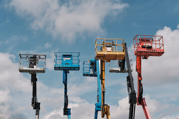 Row of colorful elevated aerial work platforms and cherry picker baskets on hydraulic booms against blue sky with white clouds. © Ekaterina