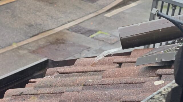 Heavy rainwater flowing from a downspout onto wet residential roof tiles. Water drainage system during a storm with a wet street below