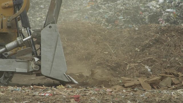 Slow motion close up shows heavy machinery pushing and leveling waste at landfill site. Dust rises as debris is moved across ground during disposal operations in landfill