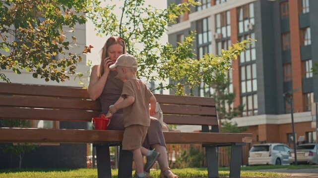 Woman sits on park bench talking while young boy stands nearby holding objects. scene takes place outdoors with trees and modern buildings in background under bright daylight