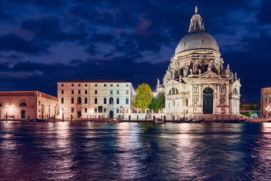 Nighttime panorama of cathedral of Santa Maria della Salute in Venice old town, Italy. Antique medieval building with dome at banks of Grand Canal. View from canal with water waves. Famous landmark..