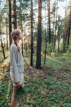 A teenage girl holds plants while standing in a summer pine forest