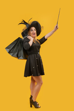 Woman dressed in a witch hat and black dress holding shopping bags and a magic wand, conjuring up the spirit of shopping deals and festive sales against a yellow background