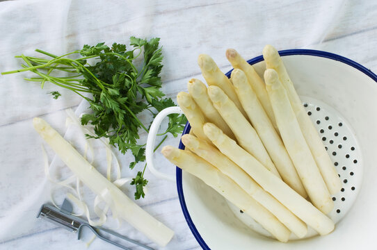 Colander of white asparagus, flat leaf parsley and peeler on white cloth